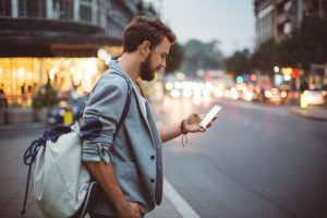 Young man on the street of big city , waiting taxi, chacking his smartphone, for news or new messages. Or looking for map instructions. Carry backpack on one shoulder. Dusk time. Casual dressed. Street lights in background.